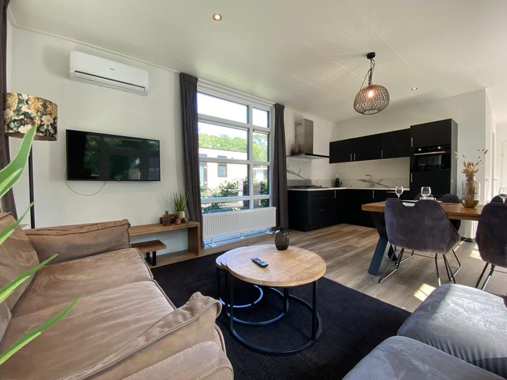Moderne woonkamer met bank, eettafel, keuken en grote ramen met uitzicht op het vakantiepark.