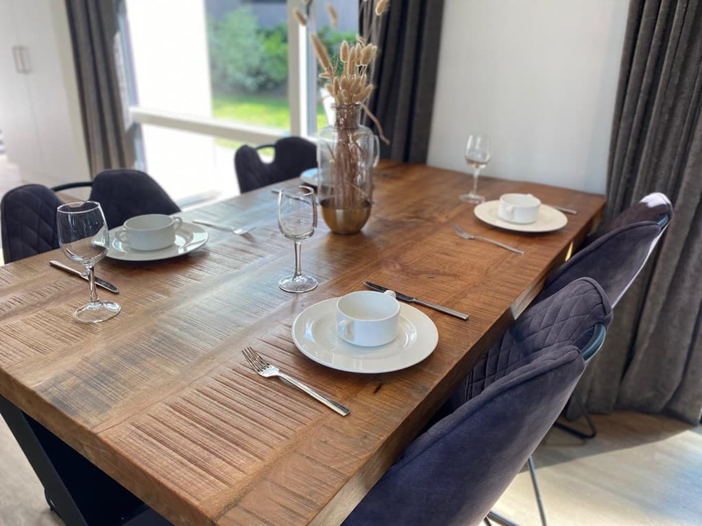Eettafel met houten blad en vier stoelen, gedekte tafel met borden en glazen, ramen met uitzicht op groen buitengebied