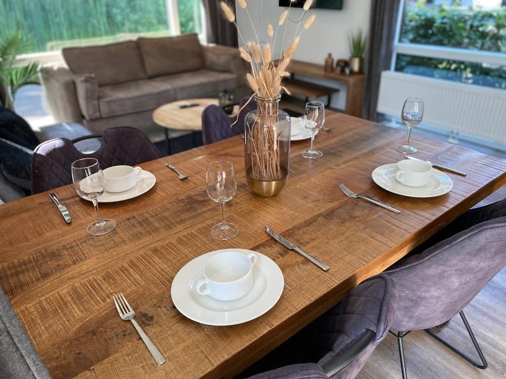 Eettafel met gedekte plaatsen, glazen, borden en een vaas met droogbloemen, in een lichte woonkamer.