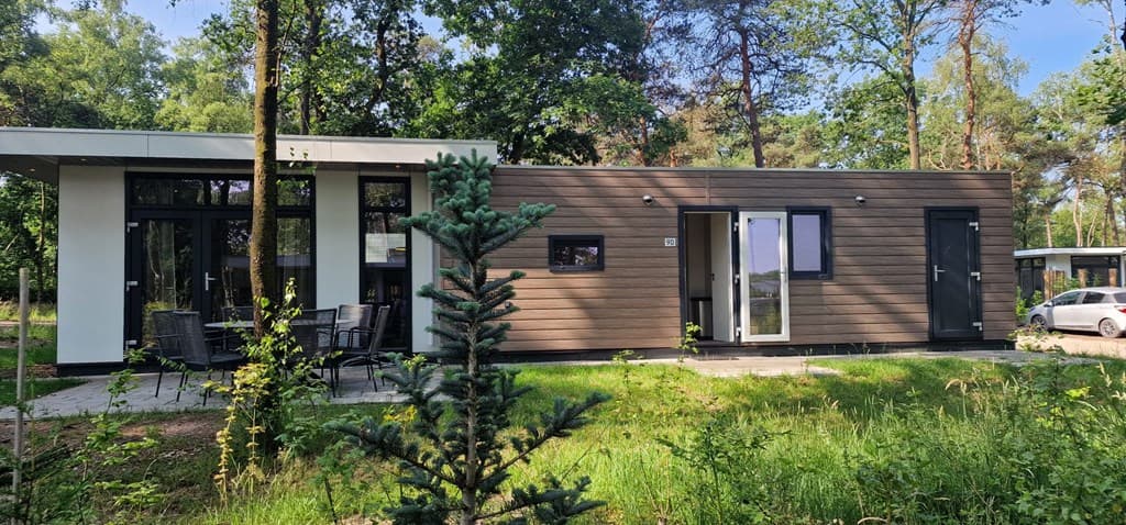 Moderne vakantiewoning met houten gevel, grote ramen, en een terras met stoelen in een bosrijke omgeving