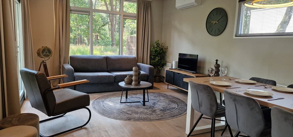 Zithoek met een grijze bank, salontafel en TV, naast een eetruimte met een tafel en stoelen, omgeven door ramen met uitzicht