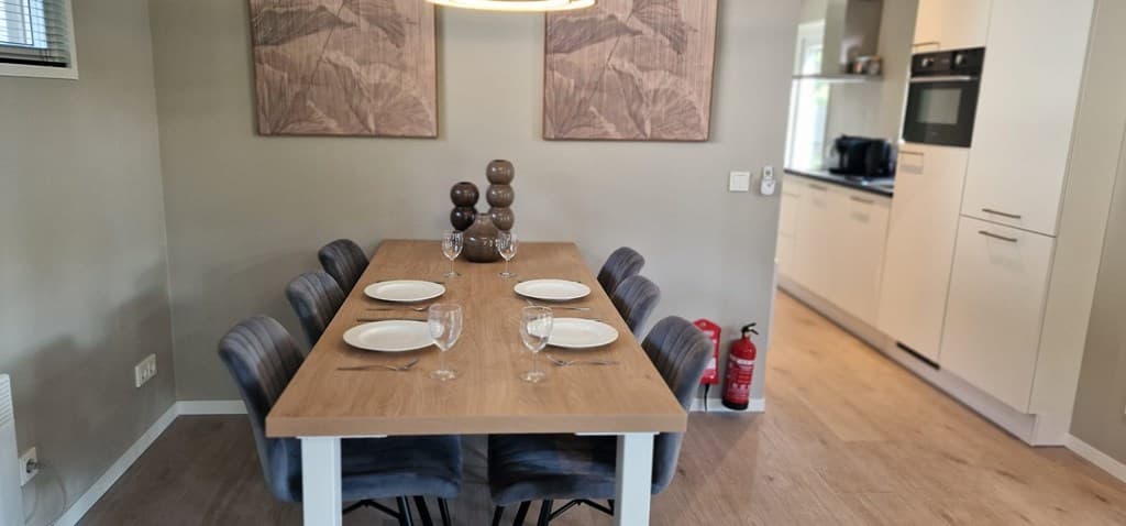 Eethoek met een lange houten tafel, zes stoelen, en decoratieve vazen; zicht op moderne keuken in achtergrond.