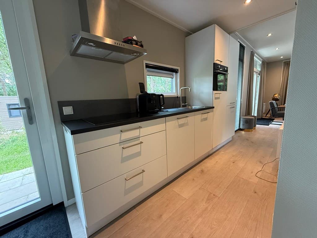 Moderne keuken met witte kasten, kookplaat en oven, naast een deur naar buiten en uitzicht op de woonkamer.