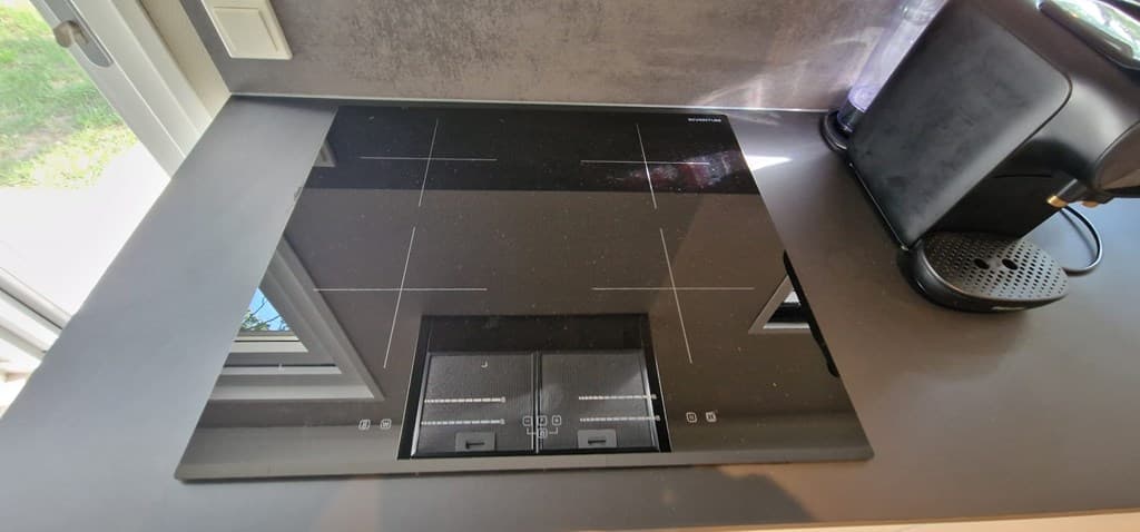 Inbouw inductiekookplaat op een keukenblad met een koffiezetapparaat ernaast en uitzicht op het buitenlicht.