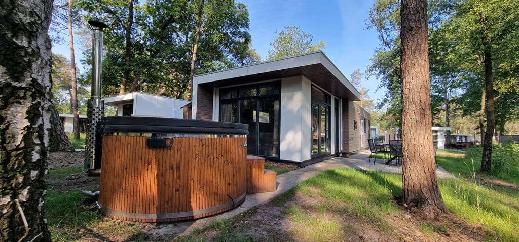 Moderne vakantiewoning met houten hot tub in een groene omgeving, omgeven door bomen en grasvelden.