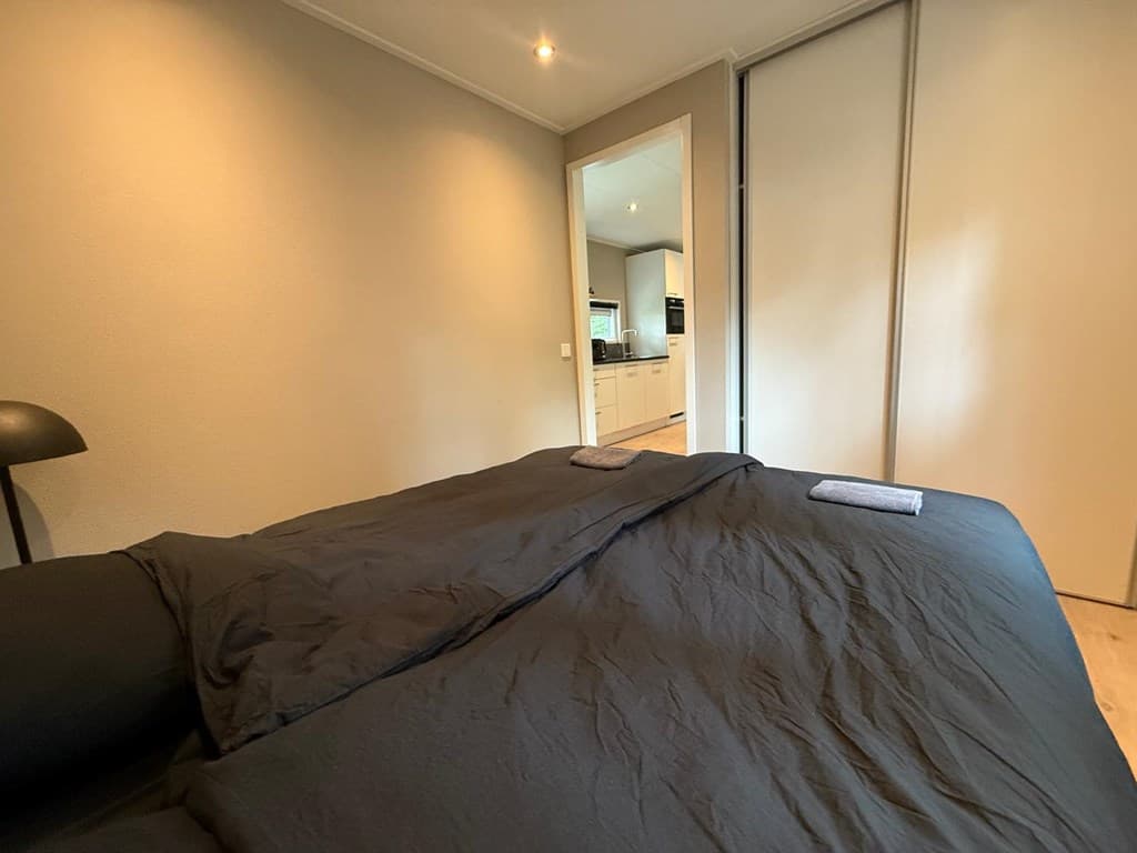 Slaapkamer met een groot bed en een deur naar de open keukenachtige ruimte met witte kasten en verlichting.