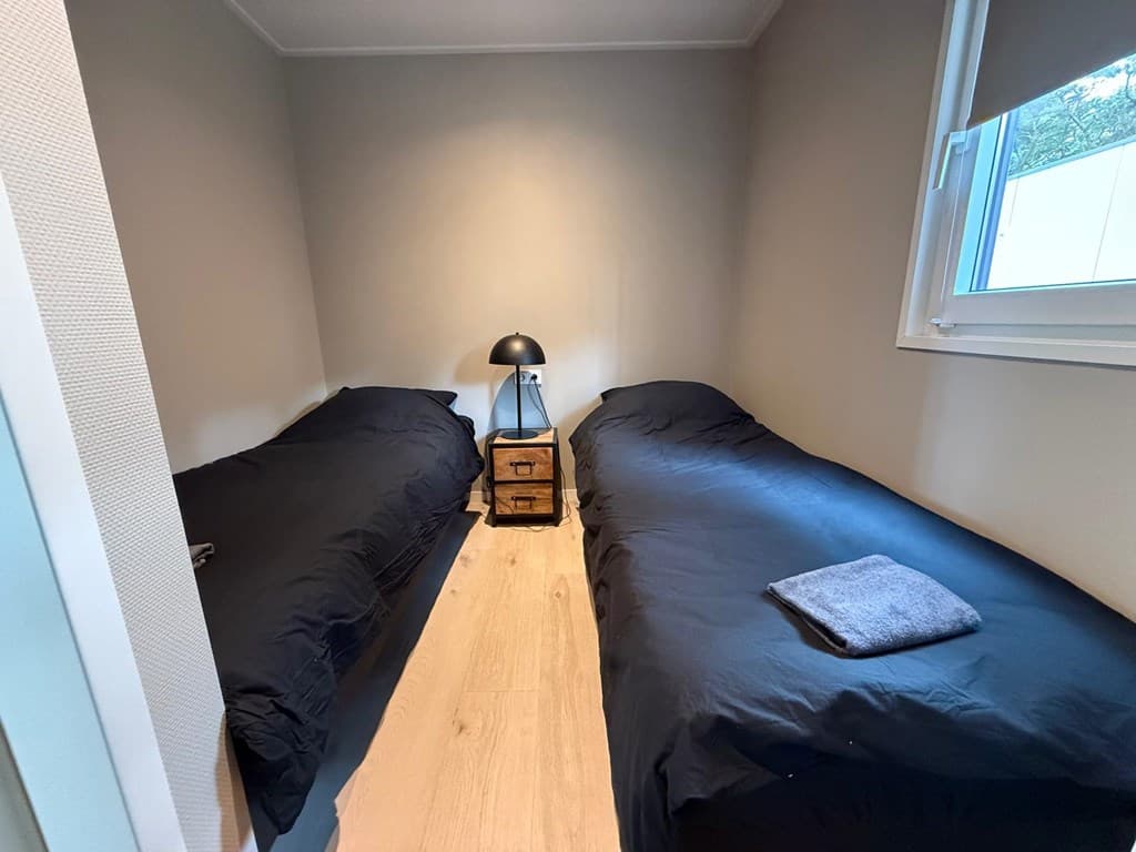 Slaapkamer met twee eenpersoonsbedden, een nachtkastje en een lamp, lichtgrijze muren en houten vloer.