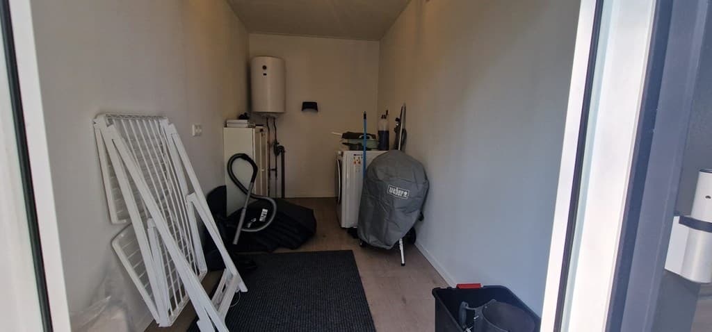 Opbergruimte met diverse accessoires zoals een wasmachine, strijkplank en tuinmeubelen in een lichte ruimte.