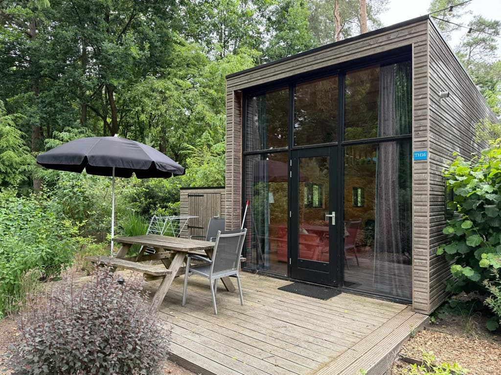 Moderne vakantiewoning met houten gevel, terras, tafel met stoelen en een parasol, omringd door groen.
