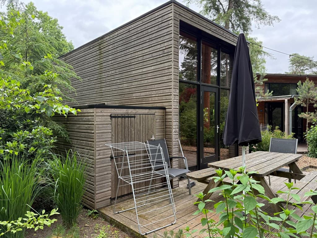 Houten vakantiehuis met grote ramen, terras met tuinmeubilair en een wasrek aan de zijkant, omringd door groen.