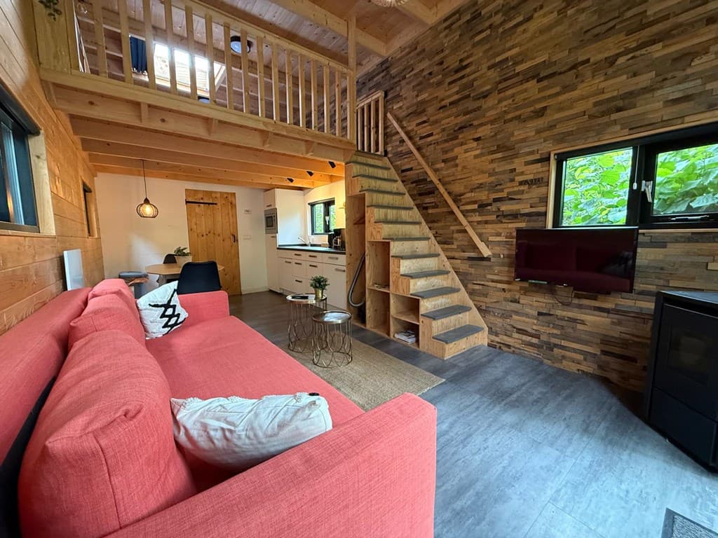 Gezellige woonkamer met een rode bank, houten accenten, open keuken en trap naar een vide met uitzicht op de natuur