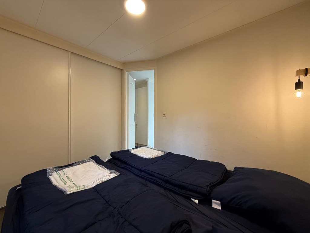 Slaapkamer met twee enkele bedden en een deur naar een andere ruimte, eenvoudige verlichting aan de wand.