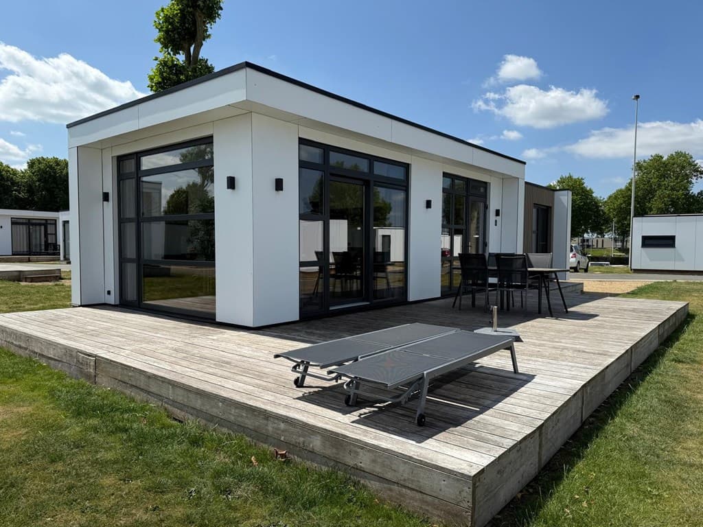 Moderne vakantiewoning met grote ramen, houten terras, ligbed en zwart tuinmeubilair in een groene omgeving