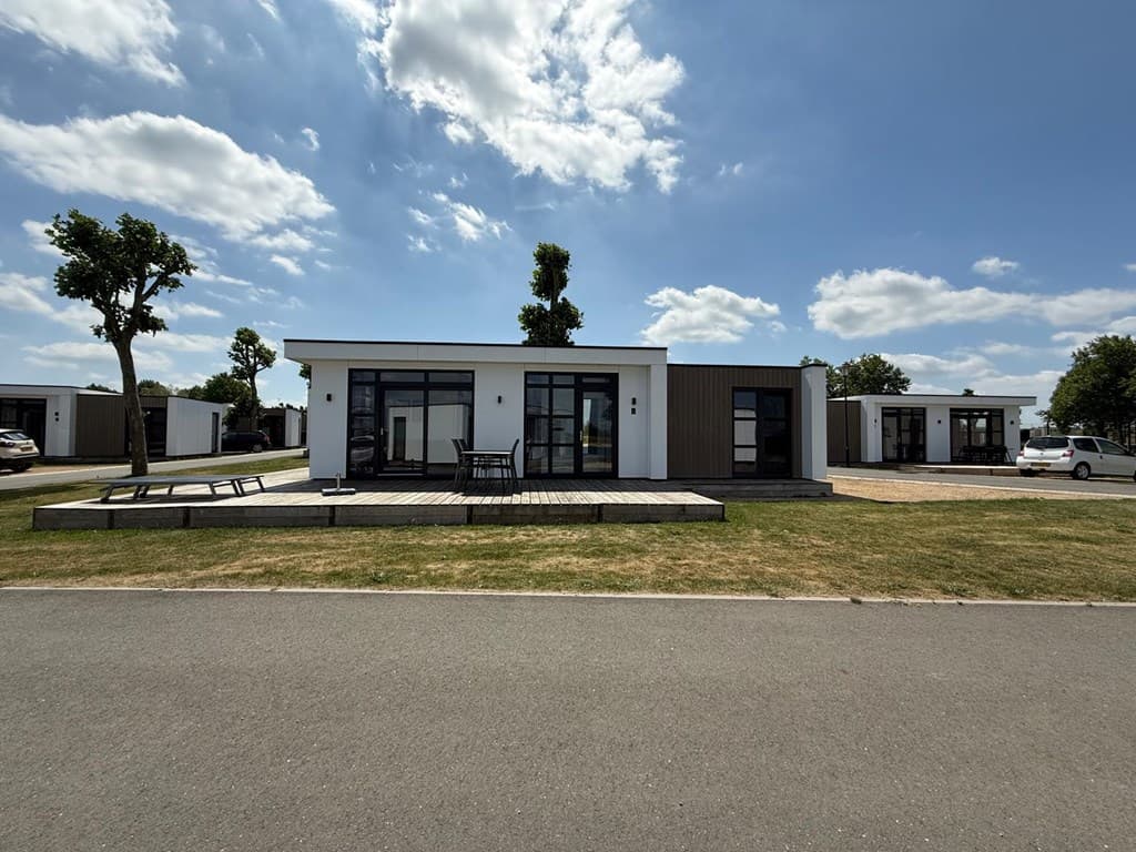 Moderne vakantiewoning met terras en grote ramen, gelegen op een grasveld tussen bomen en andere woningen.