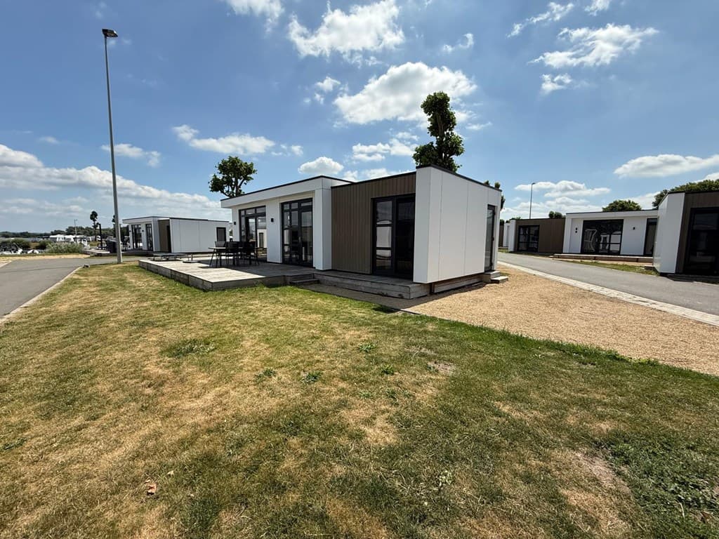 Vakantiewoning met moderne uitstraling en grote ramen, omringd door gras en verharding, met een heldere blauwe lucht.