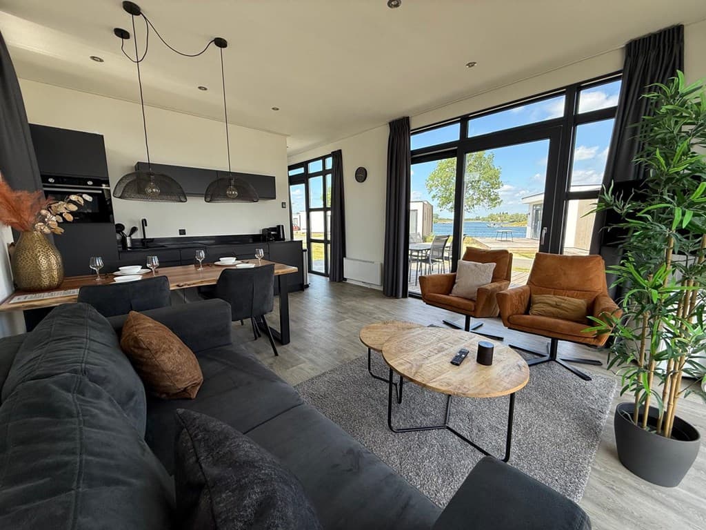 Moderne woonkamer met zithoek, eettafel en uitzicht op het water en groen door grote ramen.