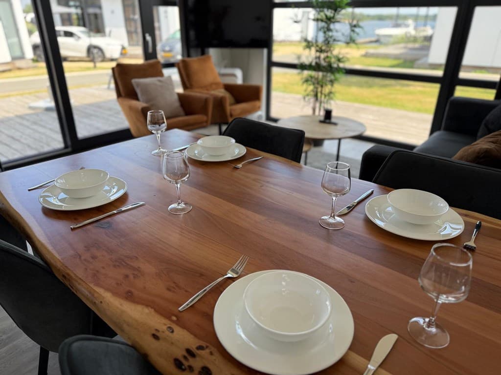 Eettafel met servies en glazen, uitzicht op de jachthaven door grote ramen, twee stoelen op de achtergrond.