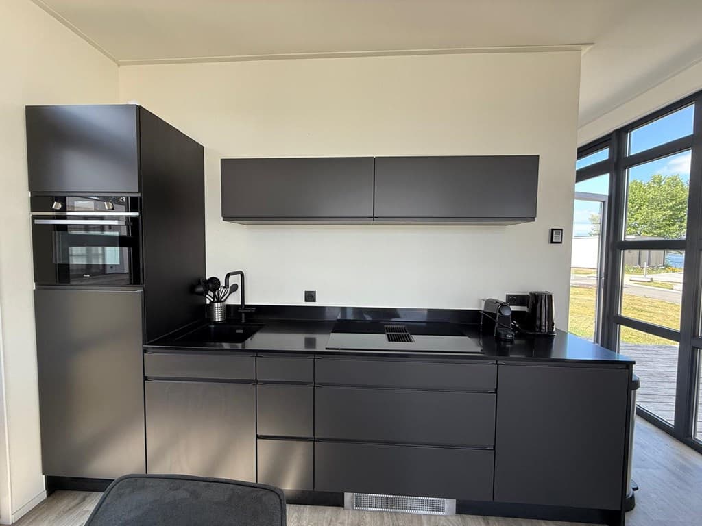 Moderne zwarte keuken met apparatuur zoals oven, koelkast en een inductiekookplaat, met veel kastruimte en een grote raampart