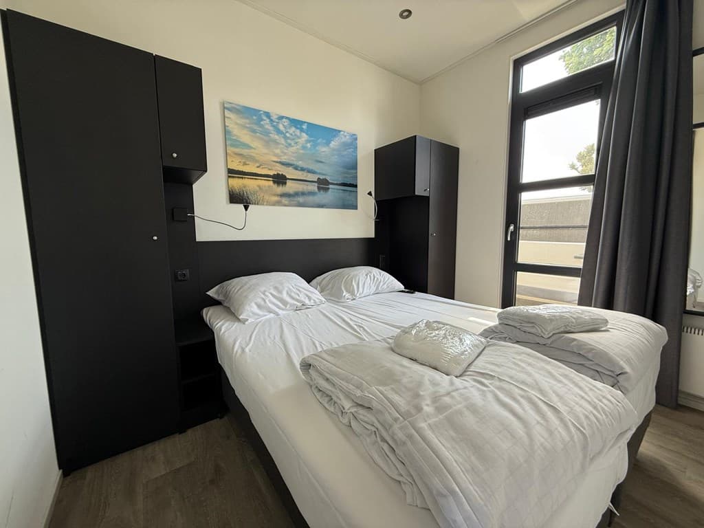 Sfeervolle slaapkamer met twee bedden, kastjes, een schilderij en grote ramen met veel natuurlijk licht.