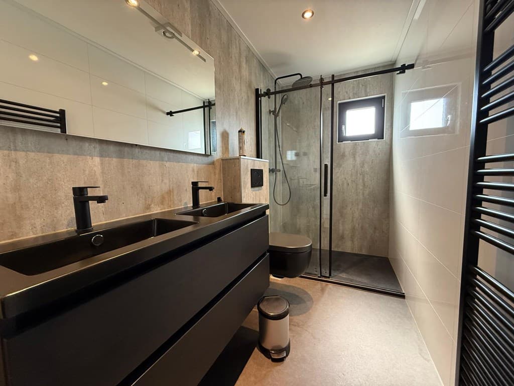 Modern badkamer met zwarte wastafel, inloopdouche en wandtoilet tegen een betonnen wand.