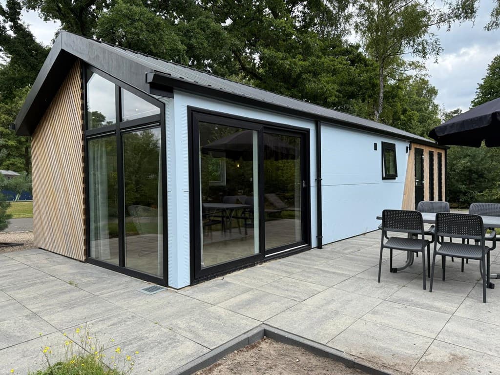 Moderne vakantiewoning met houten gevel en grote ramen, omringd door groen, met een terras en tuinmeubilair.