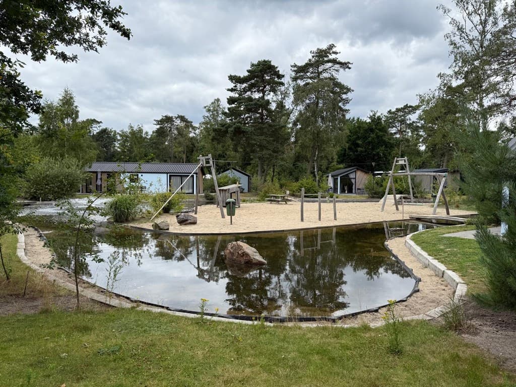 Speeltuin met zand en schommels, omringd door waterpartijen en groene bomen op EuroParcs De Hooge Veluwe.