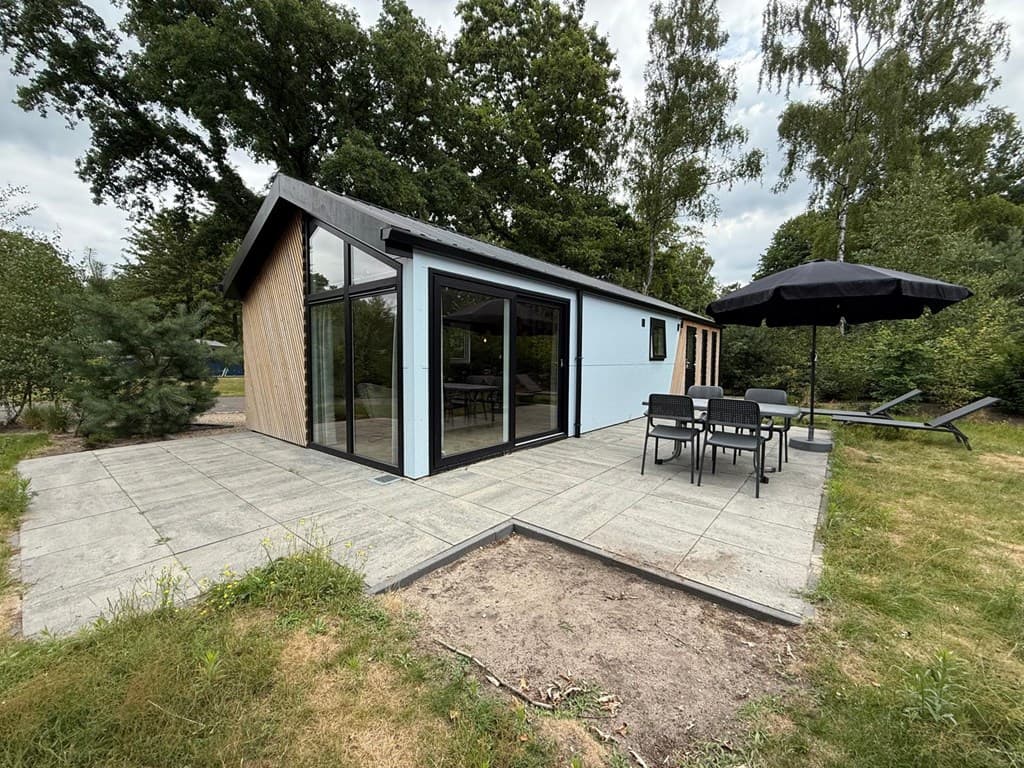 Vakantiewoning met een ruime betegelde patio, een grijze gevel en tuinmeubilair onder een parasol en ligstoel.