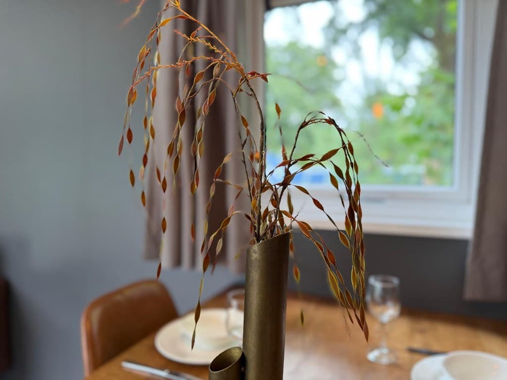 Vaasje met droogbloemen op een houten eettafel in een comfortabele woonruimte met raampartij en gordijnen.