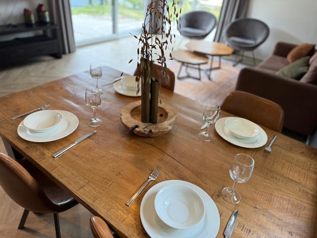 Eettafel met servies en glazen, moderne woonkamer met zithoek en grote raampartij naar buiten.
