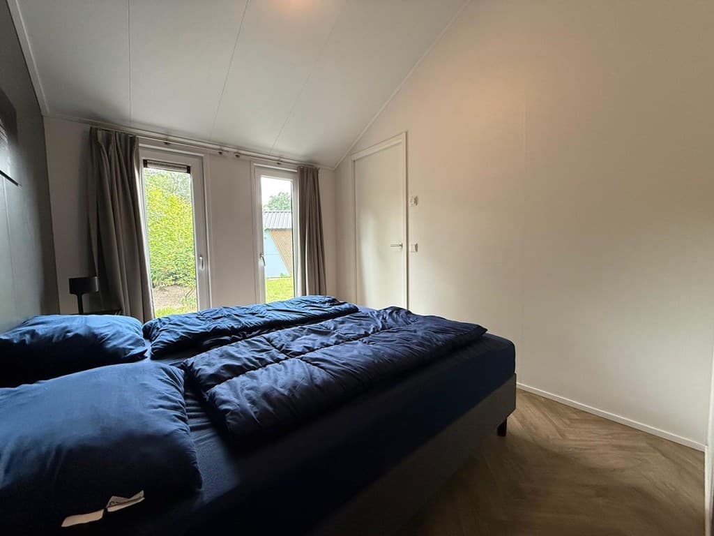 Slaapkamer met een tweepersoonsbed, gordijnen, en dubbele deuren naar de tuin. Houten vloer en eenvoudige inrichting.