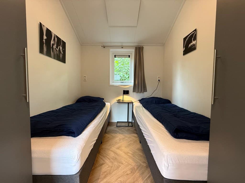 Twee eenpersoonsbedden staan in een lichte kamer met een raam en een nachtkastje tussen de bedden.
