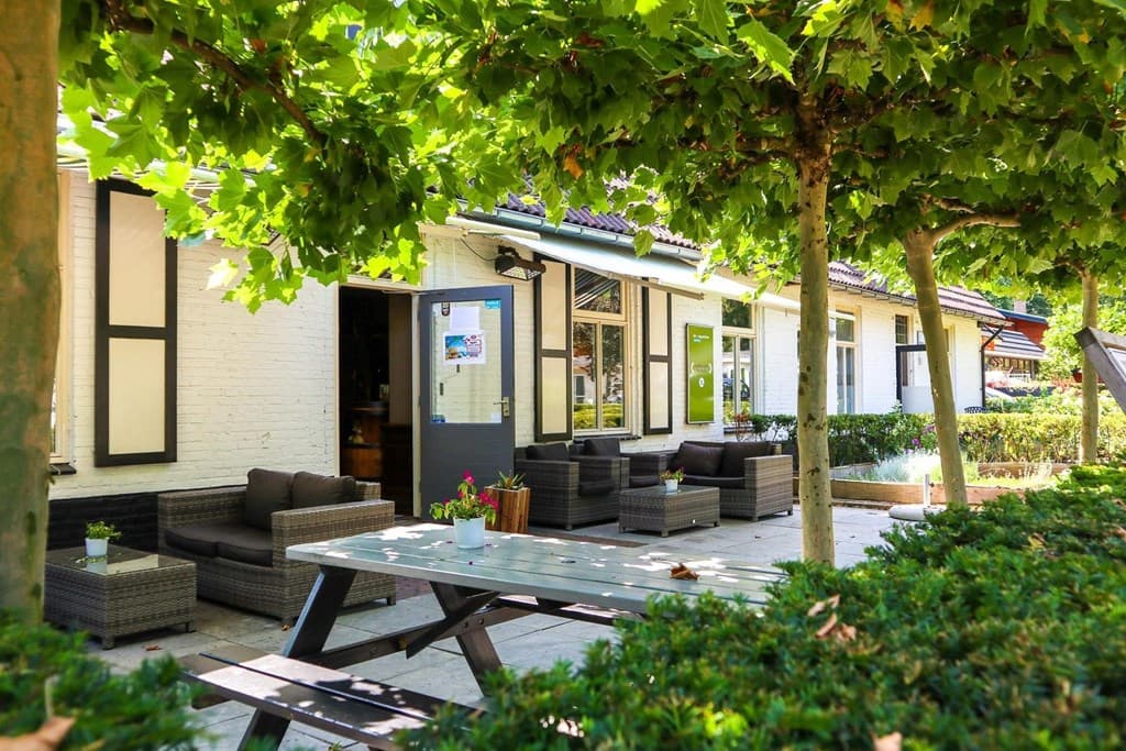 Terras met buitenmeubilair onder groene bomen, gevel van een woning met deur en grote ramen