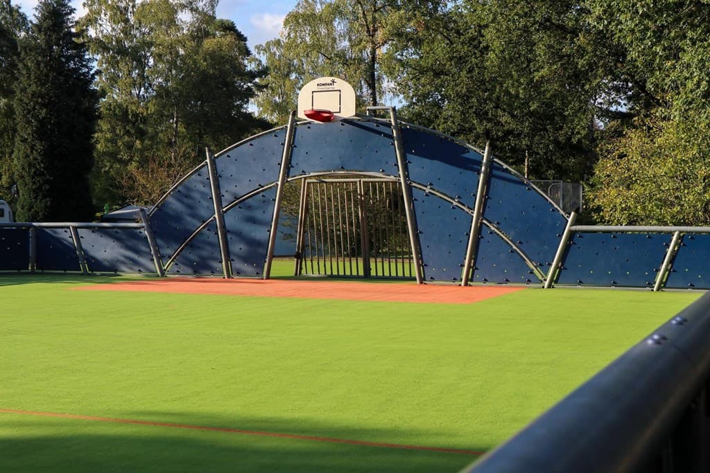 Speelveld met een blauw klimrek en een basketbalring op een groen kunstgrasveld omgeven door bomen