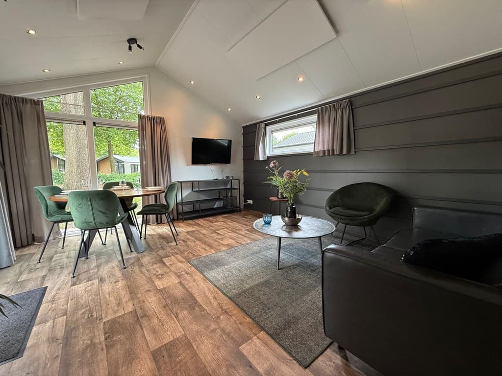 Ruime woonkamer met tafel en stoelen, een bank, fauteuils, tv en grote ramen met uitzicht op de natuur