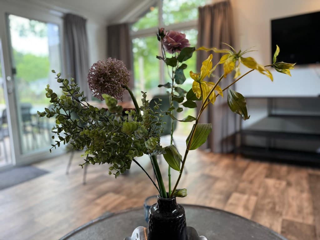 Bloemstuk met verschillende kunstplanten in een vaas op een tafel, op de achtergrond een woonkamer met grote ramen en een tel