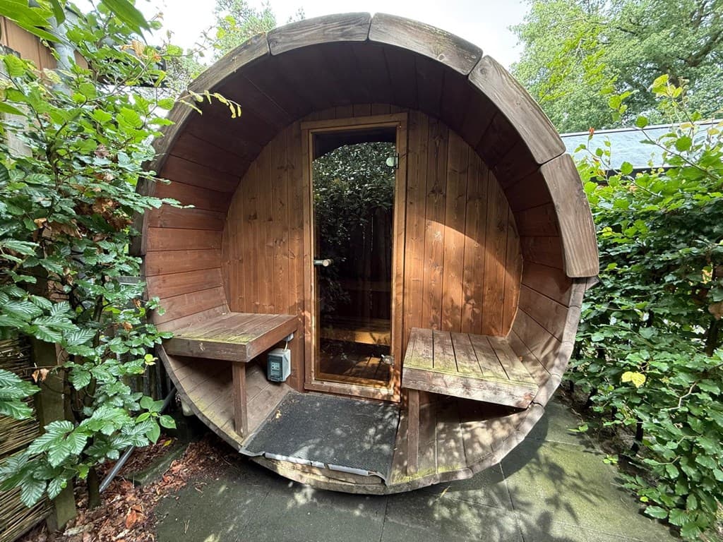 Houten sauna in een ronde vorm, omringd door groene tuinplantjes en bushaltes.