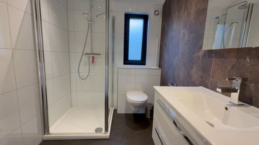 Moderne badkamer met douche, toilet en wastafel in heldere tegels en donkere vloeren.