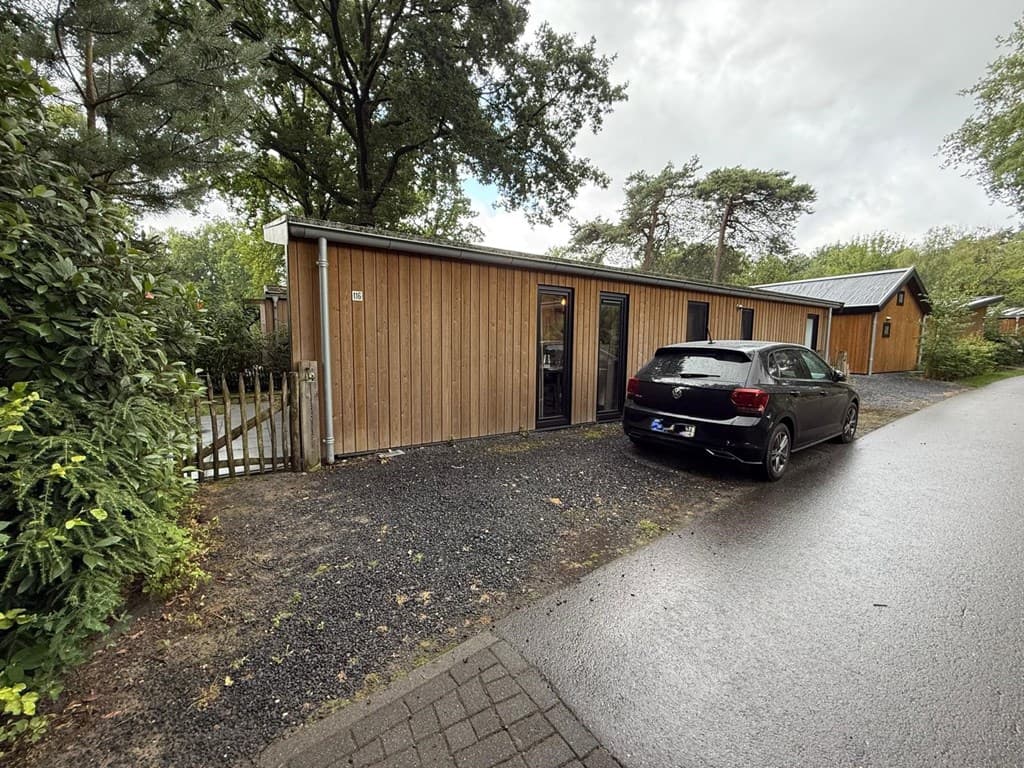 Houten vakantiewoning met een zwarte auto ernaast, omgeven door bomen en een grindpad.