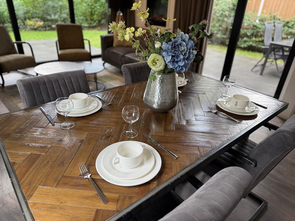 Eettafel met servies en bloemenvaas, zicht op groene tuin door grote ramen.