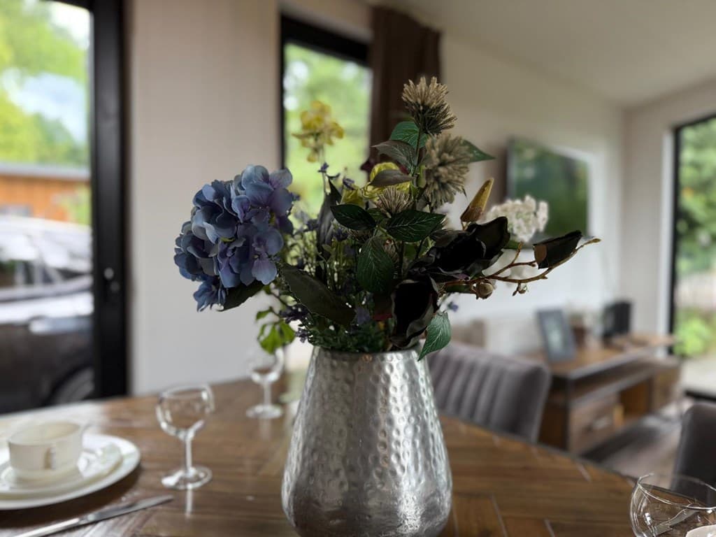 Vaas met bloemen op een houten eetkamertafel, grote ramen met uitzicht op de tuin en moderne meubels zichtbaar