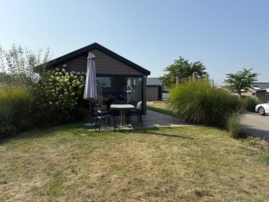 Een moderne woning met een terras, tafel en stoelen, omringd door gras en hoge planten onder een blauwe lucht