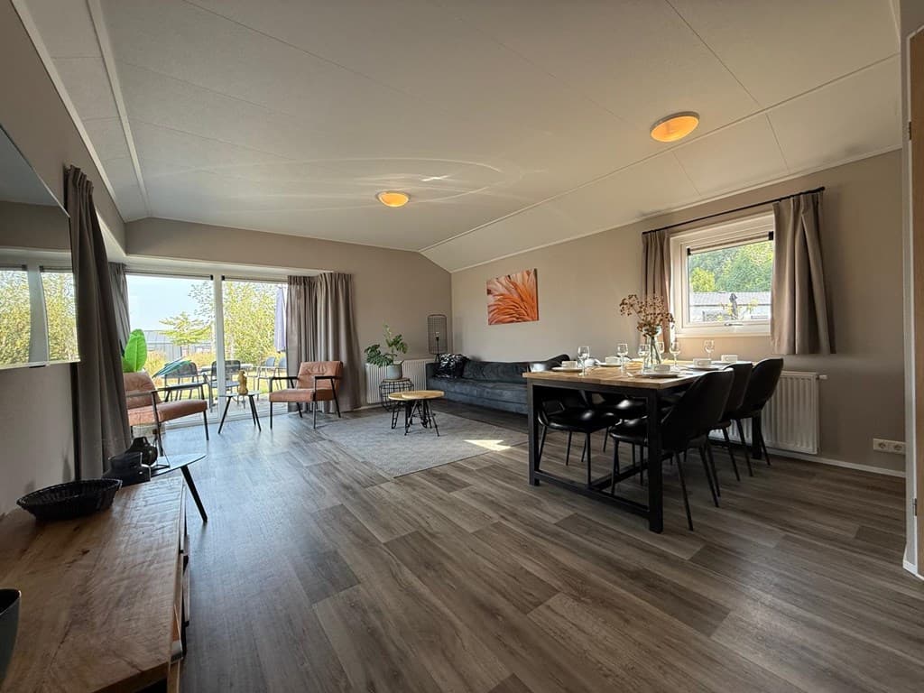 Ruime woonkamer met grote ramen, stijlvolle meubels en een eettafel met stoelen, uitzicht op de buitenruimte zichtbaar.