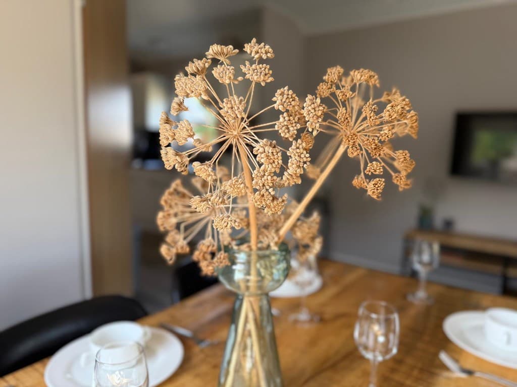 Droogbloemen in een vaas op een houten eettafel met servies en glazen op de achtergrond in een lichte ruimte