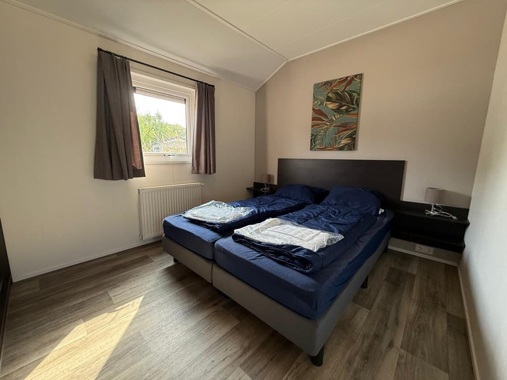 Slaapkamer met twee eenpersoonsbedden, raam met gordijnen en schilderij aan de muur. Houten vloer en nachtkastjes zichtbaar
