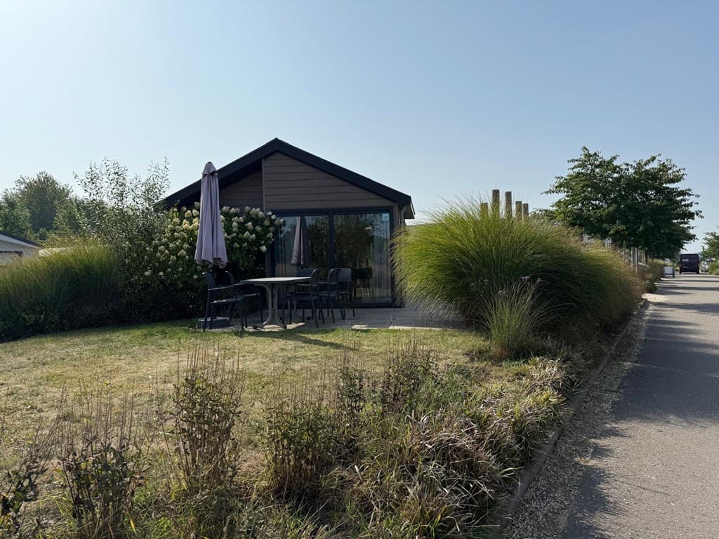 Moderne vakantiewoning met grote ramen, omringd door groene planten en een patio met tafel en stoelen