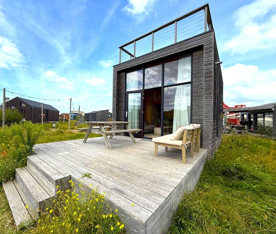 Moderne woning met grote raampartij, houten veranda, bank en tafel, omringd door gras en bloemen.