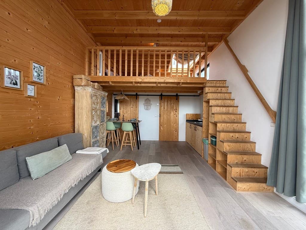 Interieur van een woning met houten wanden, trap naar vide, bank, eethoek en open keuken zichtbaar.