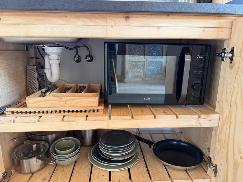 Ingebouwde keukenkast met een magnetron, schalen, pannen en bestek in houten rekken.
