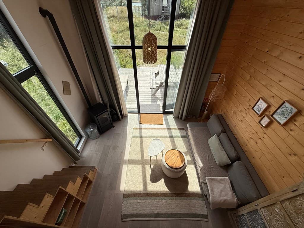 Uitzicht op de zitkamer met grote ramen, houten muren, een bank met kussens en een houten tafel op een vloerkleed.