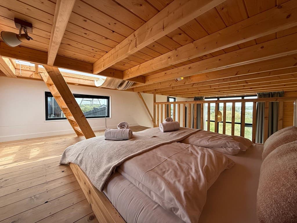 Slaapkamer met houten balken, twee opgemaakte bedden en grote ramen die licht binnenlaten.